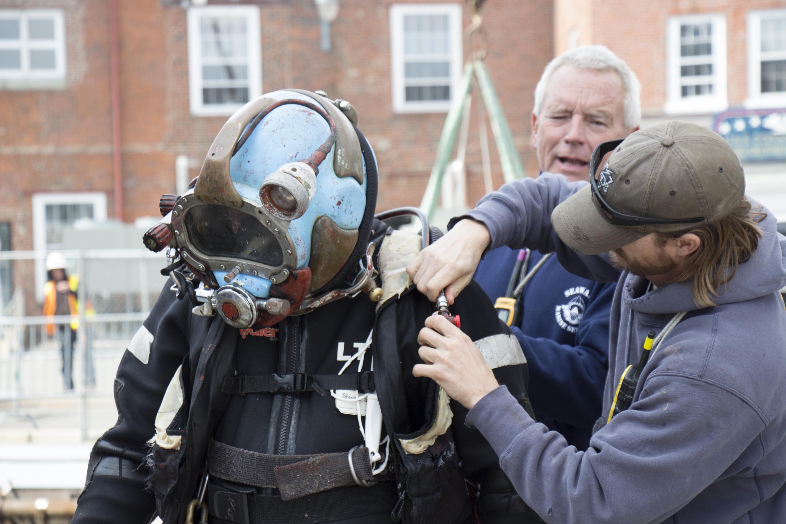Employees of Seaward Marine Services, Inc. connect air and water tubes and communication wires to a diver's helmet, April 6, 2015. [Courtesy USS Constitution Museum]